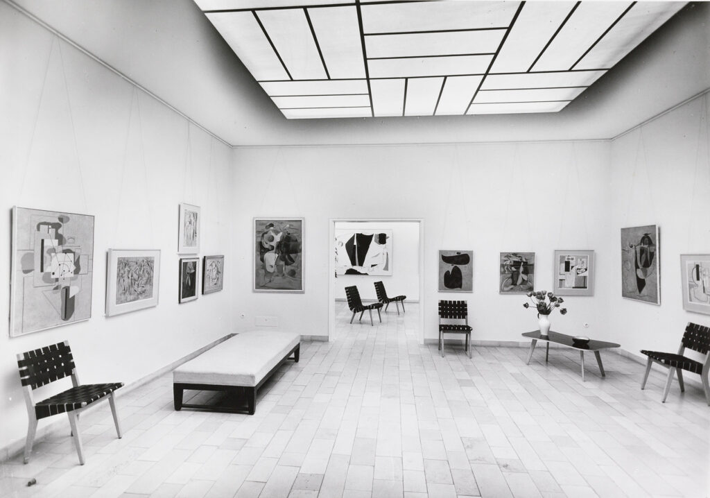 Raumansicht einer Ausstellung mit Werken an den Wänden und Möbel davor