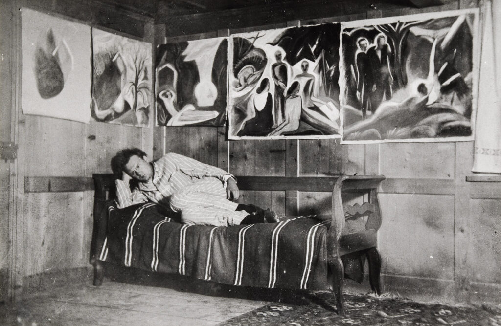 Willi Baumeister auf einer Liege vor einer Wand mit 5 Arbeiten von ihm